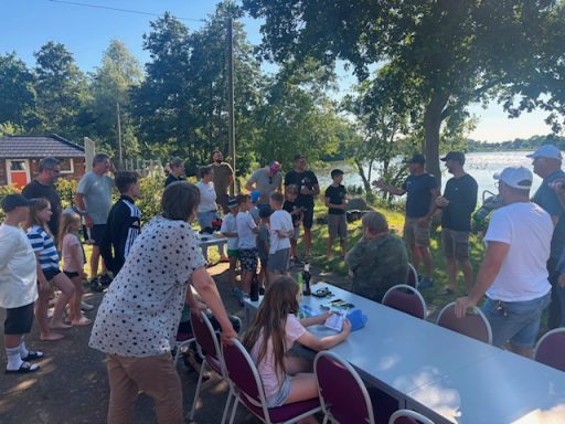 Menschen stehen um einen Tisch, im Hintergrund ein See und Bäume. Kinder und Erwachsene zusammen.