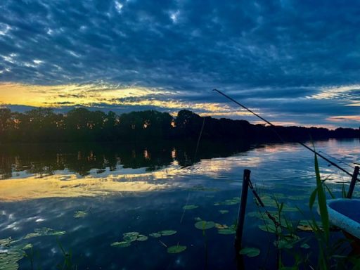 Sonnenuntergang über einem See mit Angler und spiegelndem Wasser.