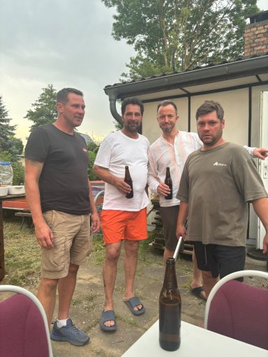 Vier Männer stehen draußen, halten Flaschen in der Hand, im Hintergrund ein Haus und Baumreihe.