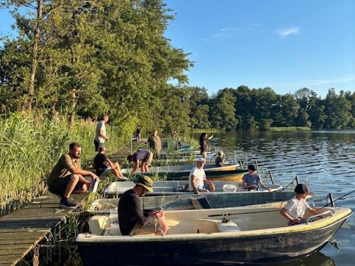 Mehrere Personen sitzen in Booten am Ufer eines ruhigen Sees bei sonnigem Wetter.