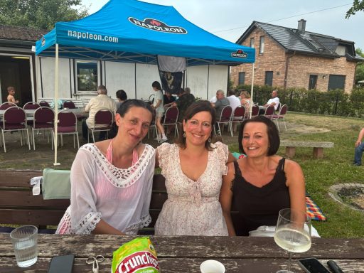 Drei Frauen lächeln im Freien vor einem Zelt, mit Gästen im Hintergrund.